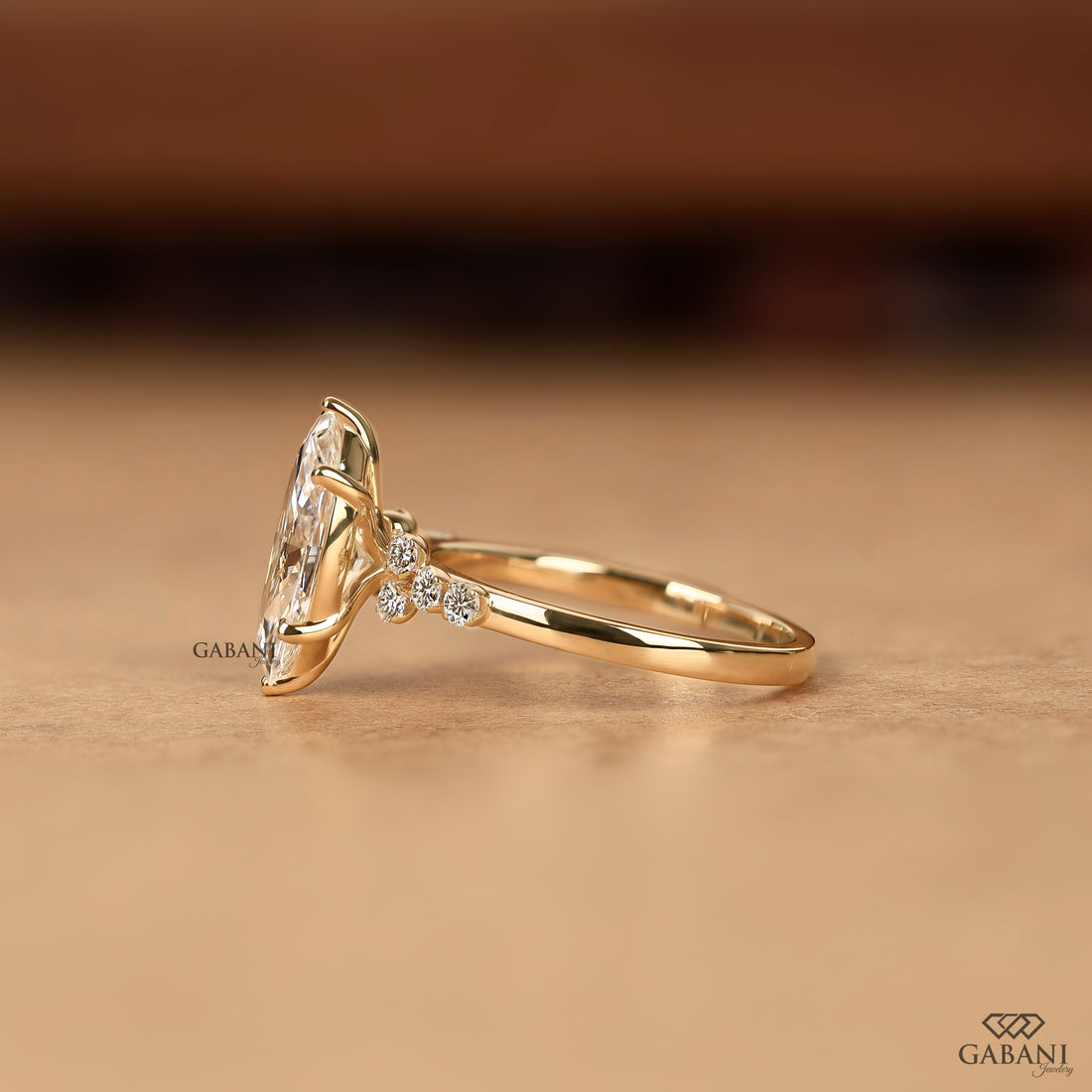 A gold ring with a large marquise clear gemstone and small accent stones on the band, photographed on a warm beige surface with a blurred brown background. The brand name GABANI appears as a watermark and in the bottom right corner.