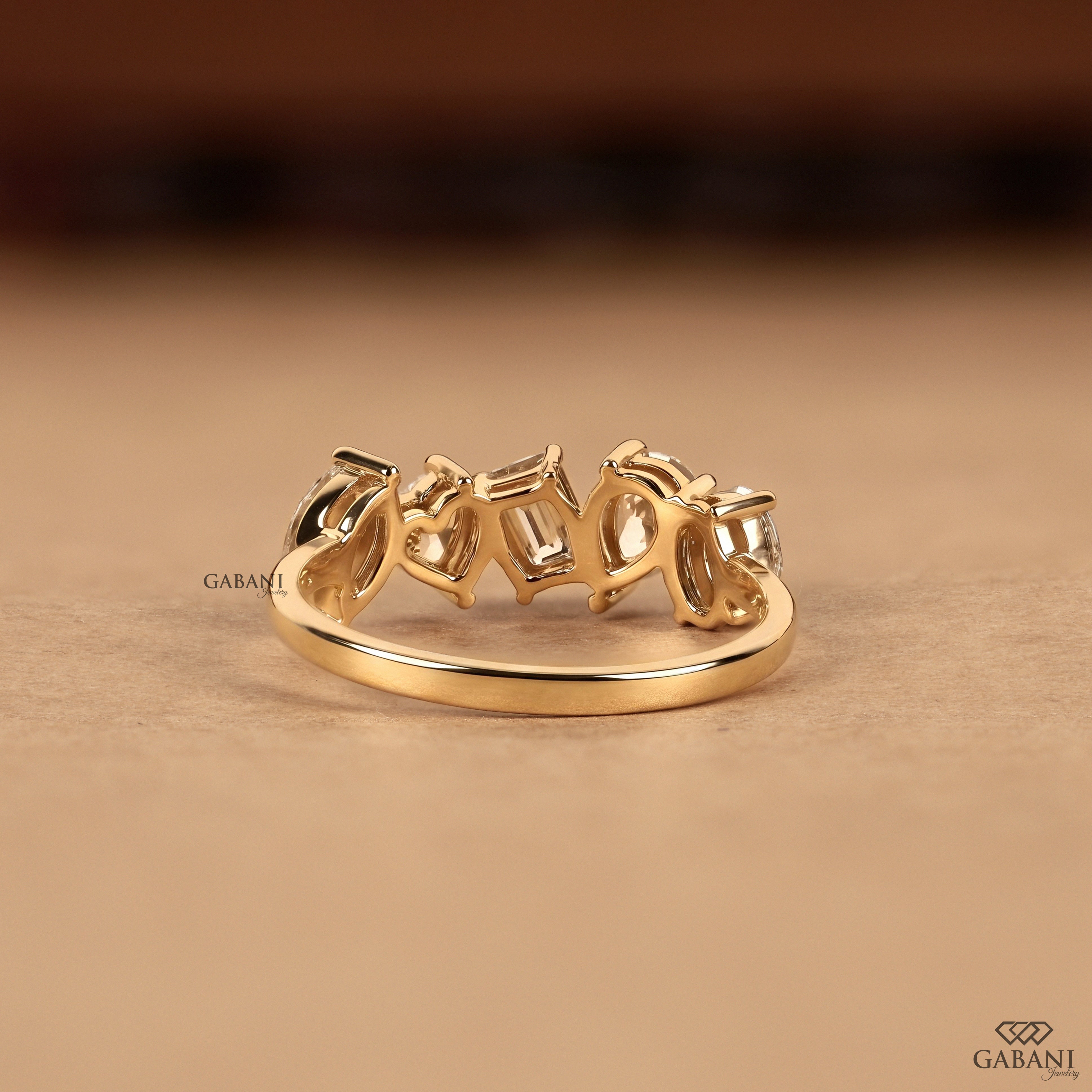 A gold ring with a row of baguette-cut clear gemstones in a prong setting, displayed on a warm beige surface with a blurred dark brown background.