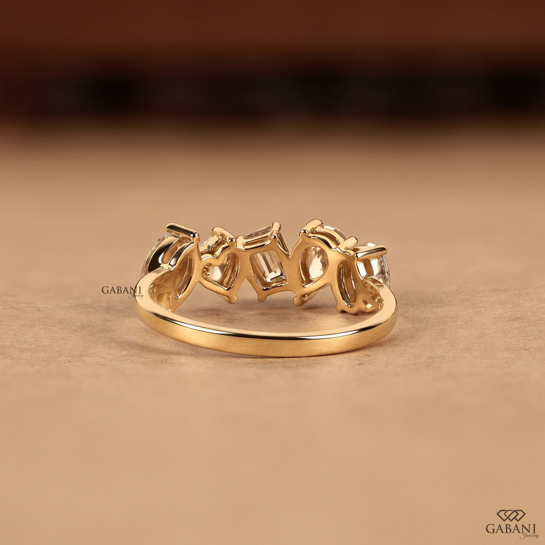 A gold ring with a row of baguette-cut clear gemstones in a prong setting, displayed on a warm beige surface with a blurred dark brown background.
