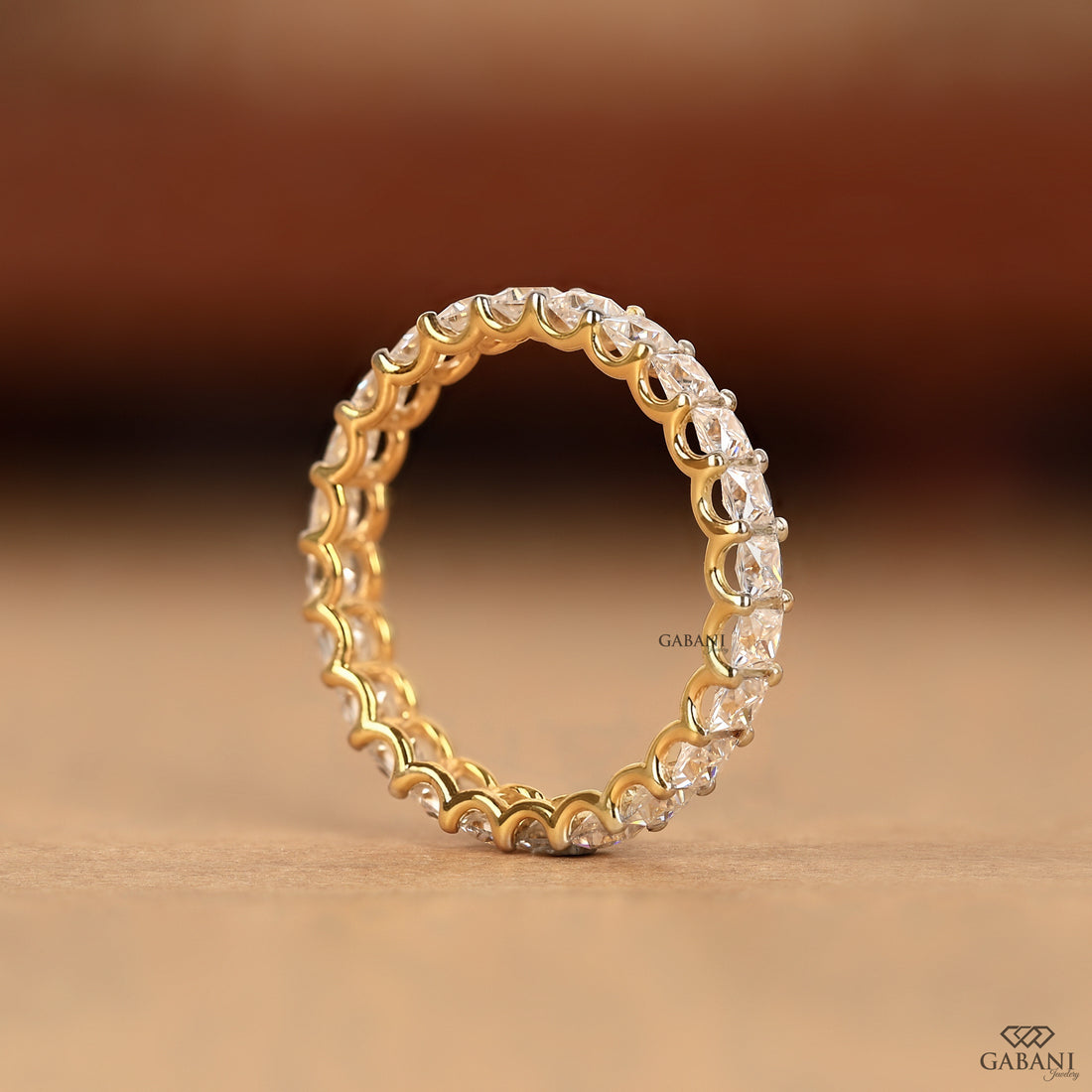 A gold eternity ring set with clear marquise-cut gemstones, standing upright on a neutral beige surface with a warm brown blurred background. The brand name 'GABANI' is visible as a watermark and logo in the lower right corner.