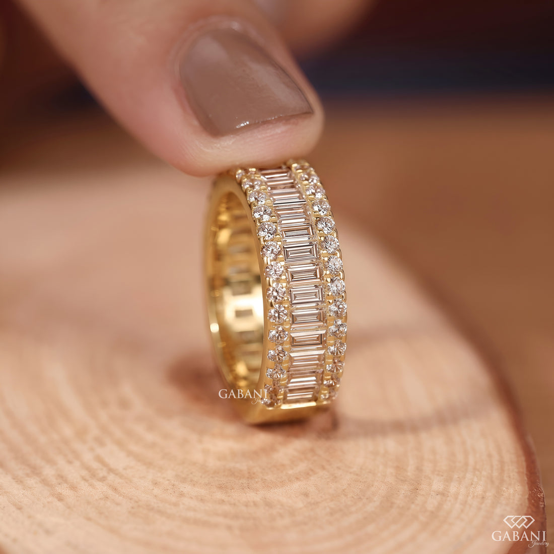Holding a wide yellow gold wedding band with baguette and round lab grown diamonds between fingers